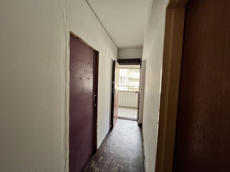 A narrow apartment hallway featuring parquet flooring and access to a security gate, showing signs of wear and requiring cosmetic updates.