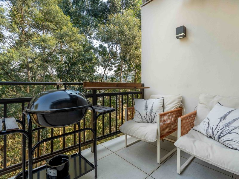 A private balcony featuring tiled flooring, a metal balustrade, and a wall-mounted light fixture, overlooking a lush green treed area.