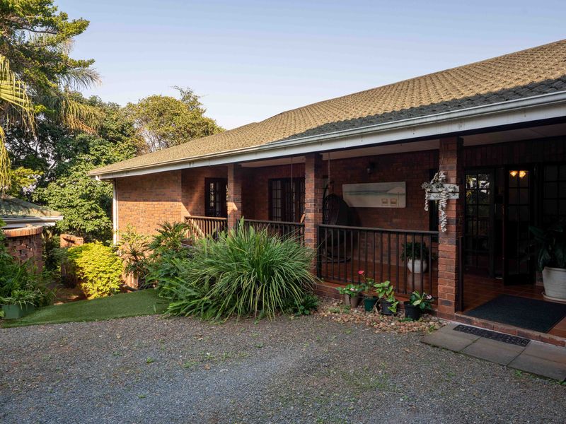 A charming brick-faced home featuring a covered front patio with tiled flooring, metal railings, and a gravel driveway entrance.
