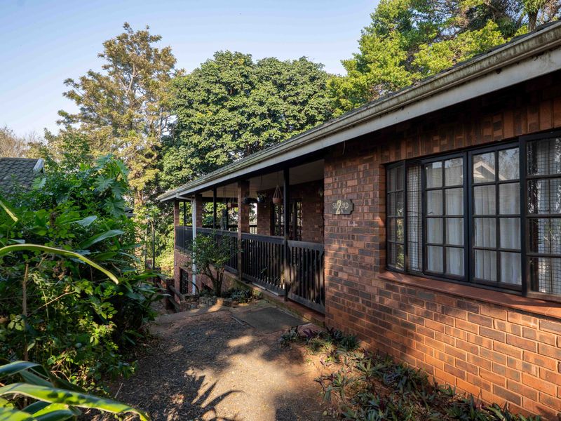A view of the front brick facade featuring a covered veranda with railings and secure window bars, surrounded by mature garden greenery.