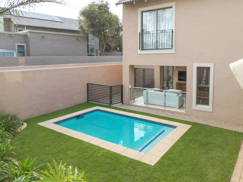 Rear exterior view featuring a swimming pool, artificial lawn, paved patio area, and a covered outdoor entertainment space with a built-in braai.