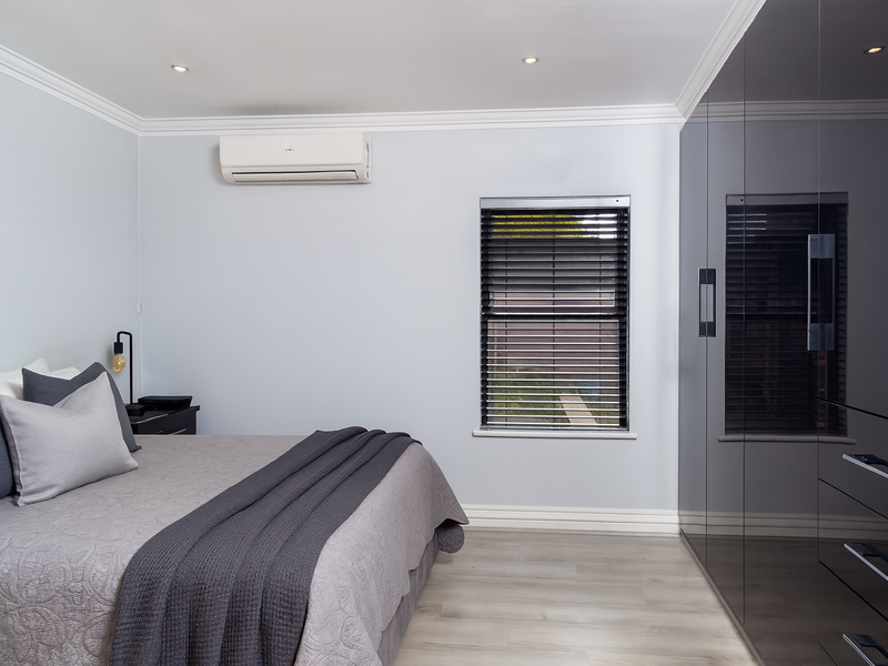 Modern bedroom featuring high-gloss built-in wardrobes, split-system air conditioning, recessed downlights, and neutral finishes with timber-look flooring.