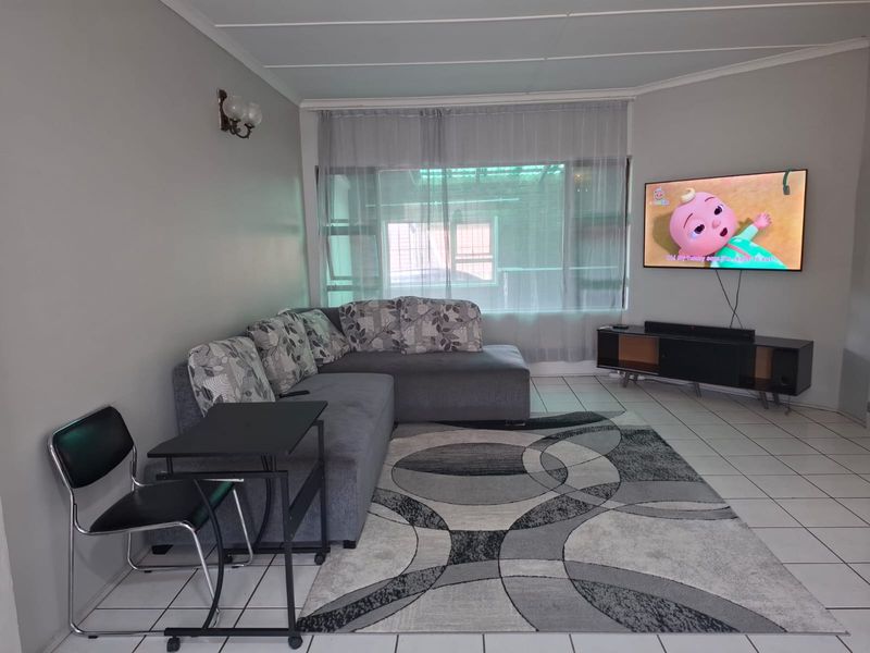 A living room featuring neutral wall tones, tiled flooring, and a large window, providing a functional and well-lit space for relaxation.