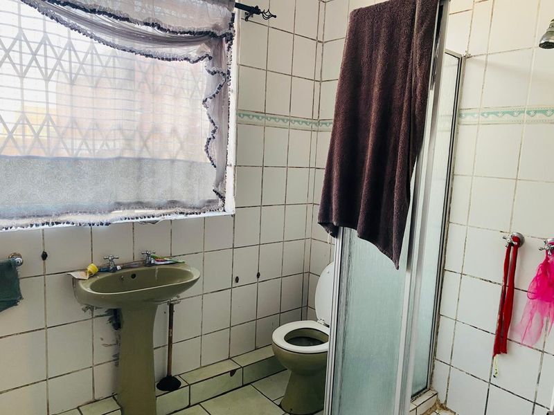A dated bathroom featuring a pedestal basin, toilet, and shower enclosure with full-height wall tiling and a security-fitted window.
