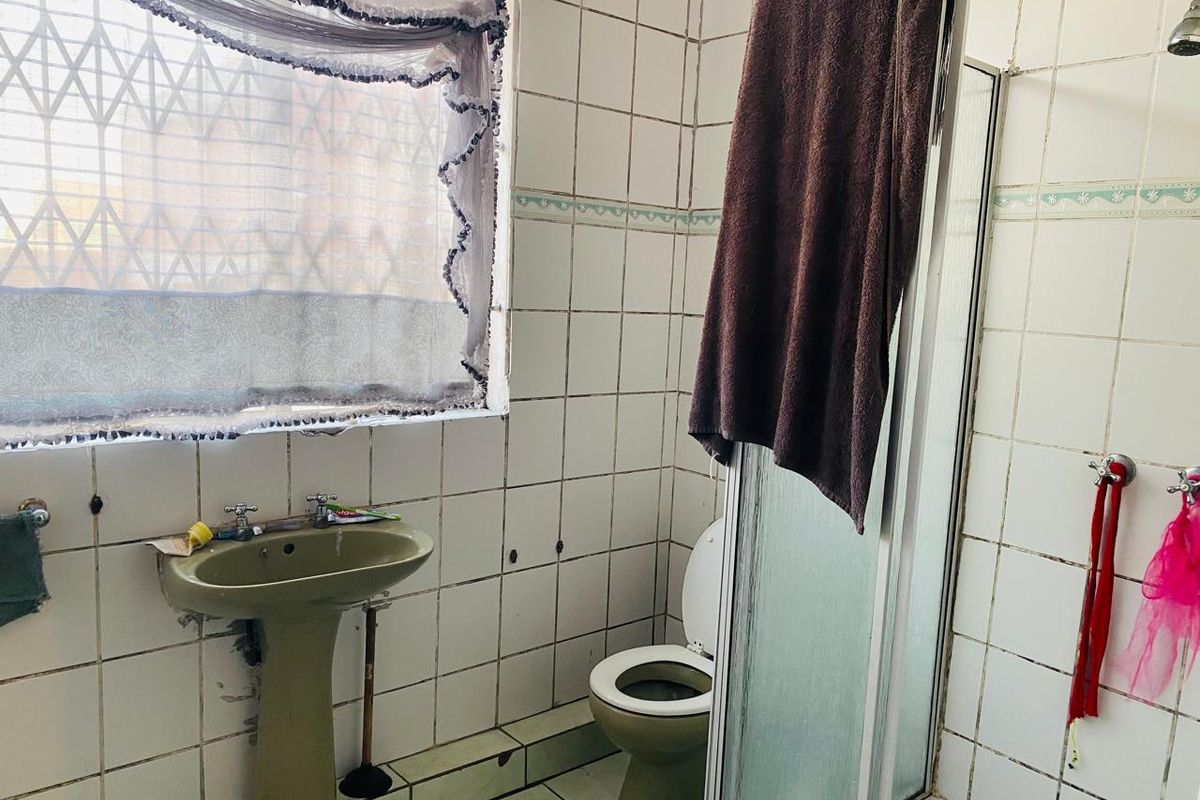A dated bathroom featuring a pedestal basin, toilet, and shower enclosure with original ceramic wall tiling and a window security gate.