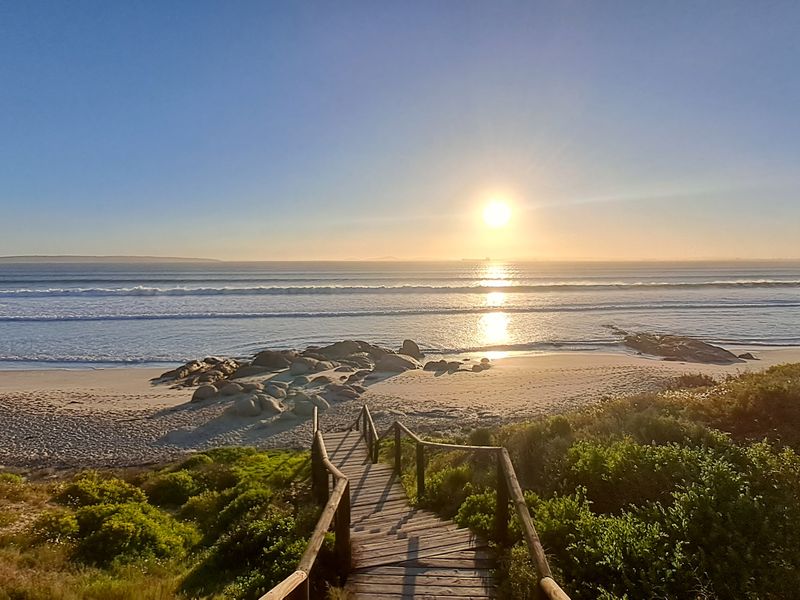 A scenic view of the ocean at sunset, featuring a wooden boardwalk leading down to the sandy beach and rocky coastline.