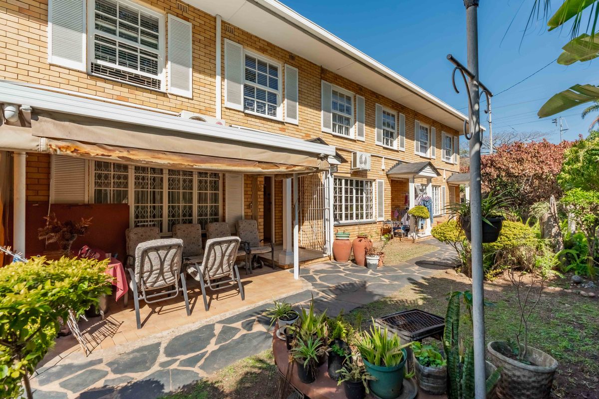 Rear view of a brick townhouse featuring a paved patio area with a retractable awning, security gates, and established garden surroundings.