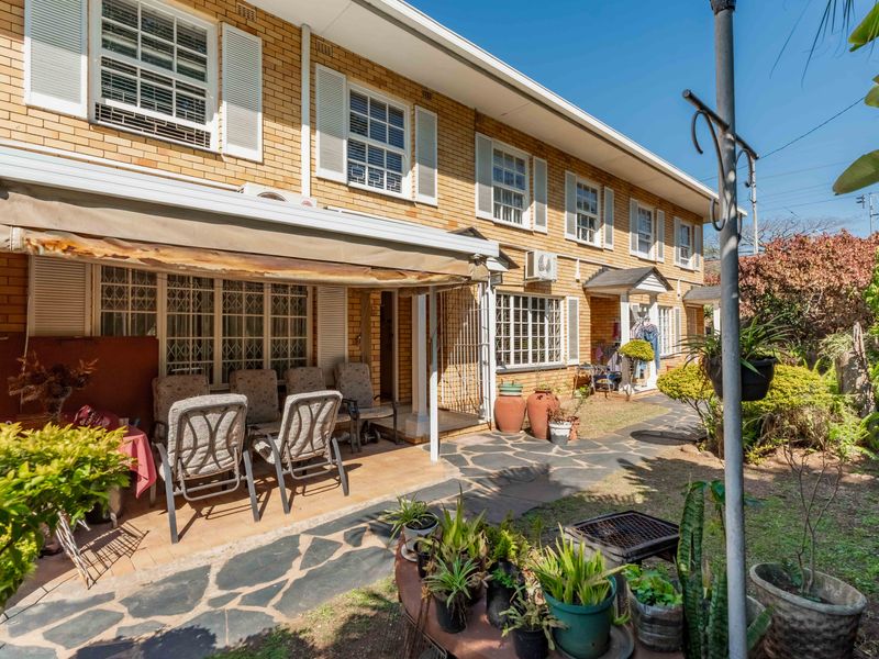 Rear view of a brick townhouse featuring a paved patio area with a retractable awning, security gates, and established garden surroundings.