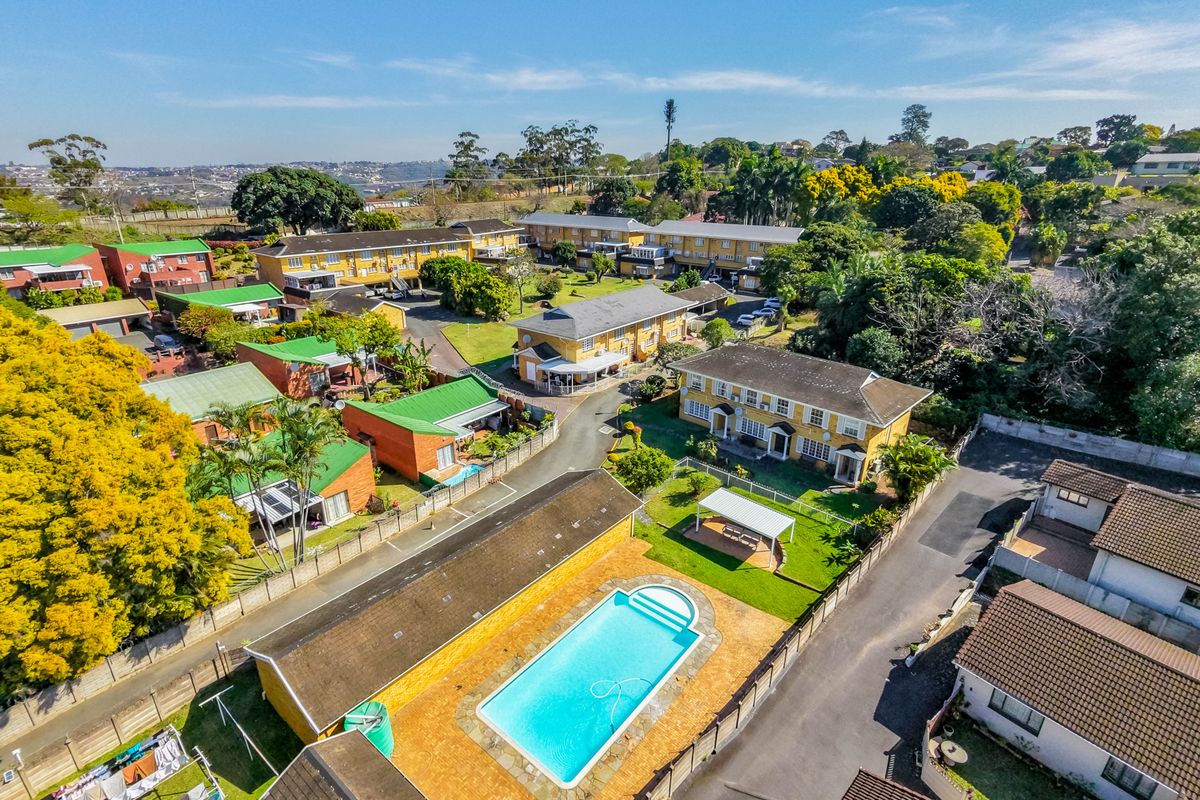 Aerial view of a townhouse complex featuring a communal swimming pool, landscaped gardens, paved walkways, and multiple residential units with tiled roofs.