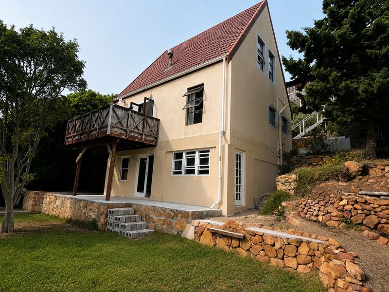 Rear view of a two-story house featuring a timber balcony, stone retaining walls, and a tiled roof set within a landscaped garden.