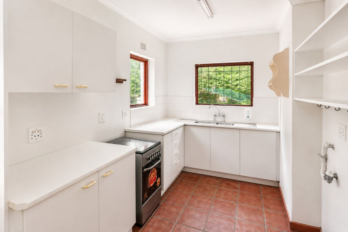 A functional, clean kitchen featuring white cabinetry, a stainless steel sink, a freestanding stove, and terracotta-style floor tiles with ample natural light.