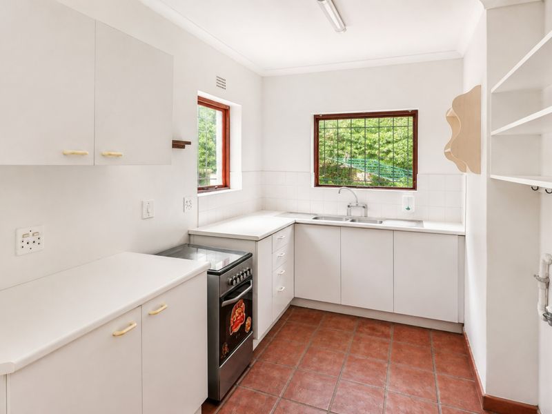 A functional, clean kitchen featuring white cabinetry, a stainless steel sink, a freestanding stove, and terracotta-style floor tiles with ample natural light.