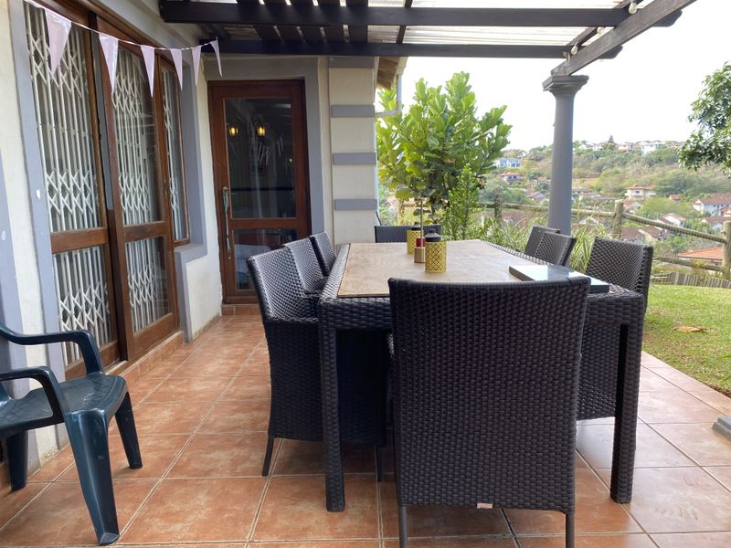 A covered outdoor patio featuring tiled flooring, a timber pergola structure, and sliding glass doors with security gates, overlooking a scenic landscape.