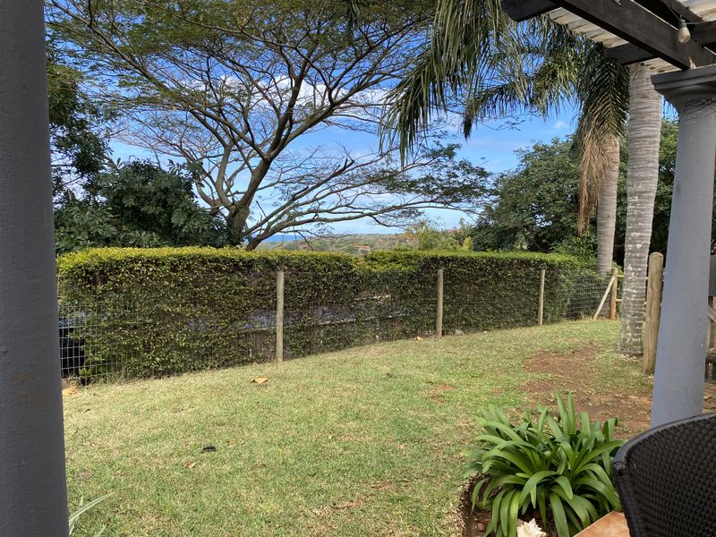 A covered patio area overlooking a private garden with established trees, a manicured hedge, and wire mesh perimeter fencing.