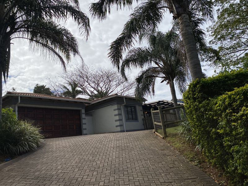 Front view of the property featuring a paved driveway, double garage with timber doors, and established garden surroundings.