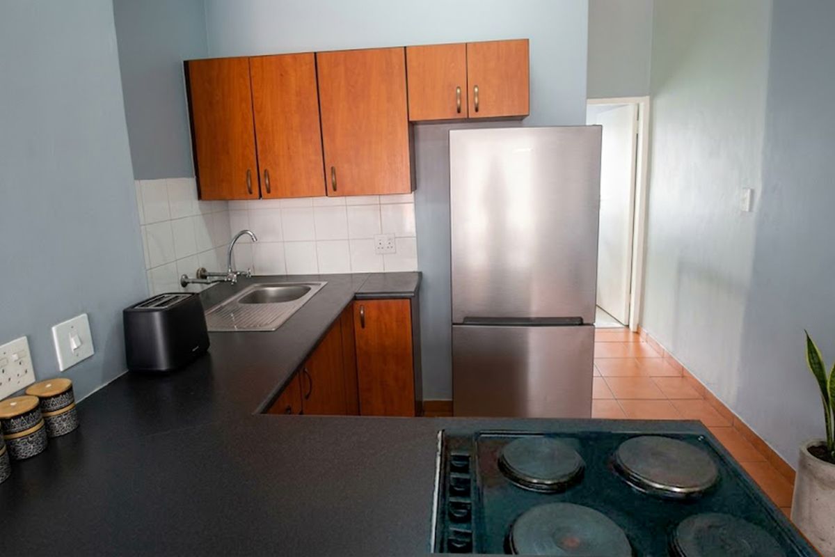 A functional kitchen featuring wood-finish cabinetry, dark laminate countertops, a stainless steel sink, and a built-in electric hob with tiled splashbacks.
