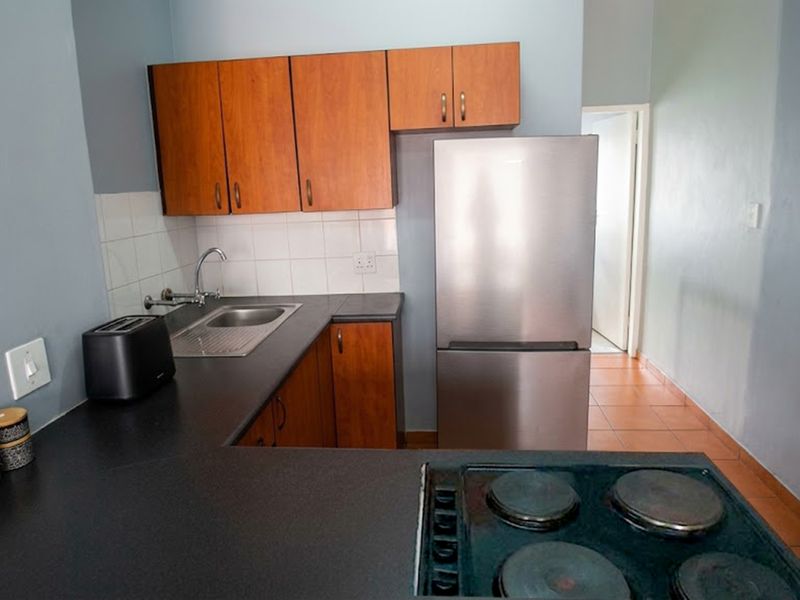 A functional kitchen featuring wood-finish cabinetry, dark laminate countertops, a stainless steel sink, and a built-in electric hob with tiled splashbacks.