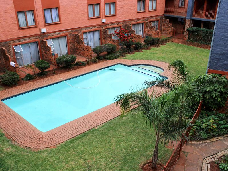 An elevated view of a communal swimming pool area set within a landscaped garden, surrounded by brick apartment building facades.