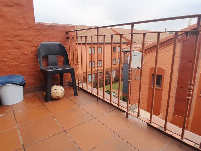 An open-air balcony featuring tiled flooring and a metal balustrade, overlooking the apartment complex grounds.
