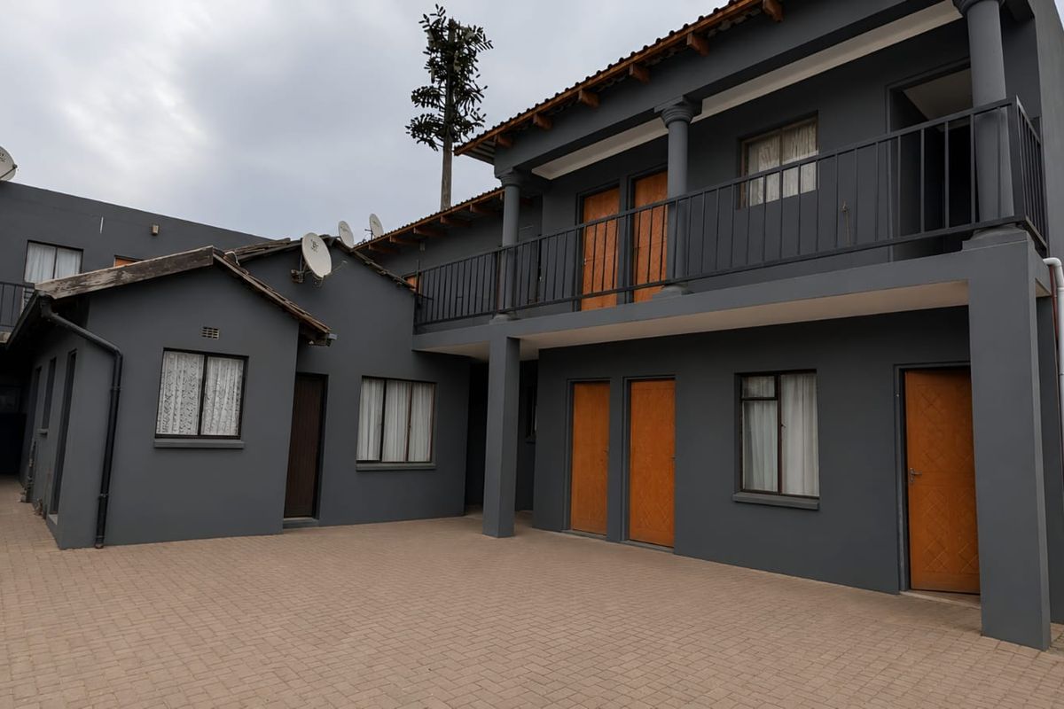 Rear exterior view of a multi-level apartment complex featuring a paved courtyard, balcony with metal railings, and external access stairs.
