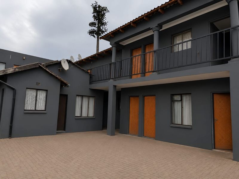Rear exterior view of a multi-level apartment complex featuring a paved courtyard, balcony with metal railings, and external access stairs.