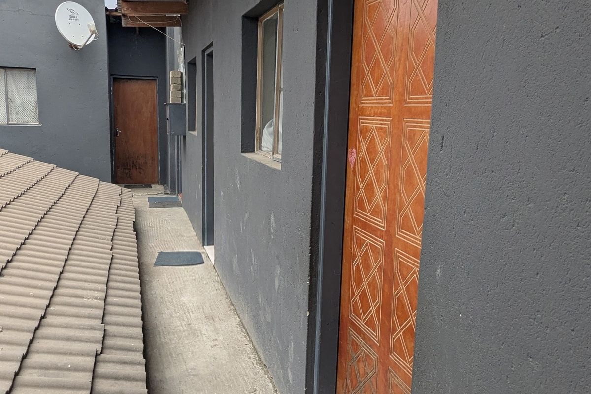 Exterior walkway featuring a concrete path, dark painted walls, and wooden doors, with exposed timber rafters and a mix of tiled and corrugated roofing.