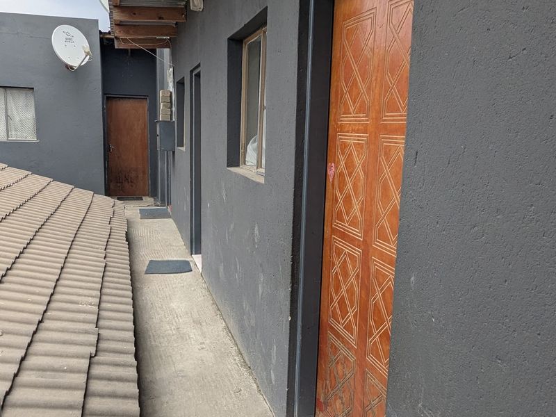 Exterior walkway featuring a concrete path, dark painted walls, and wooden doors, with exposed timber rafters and a mix of tiled and corrugated roofing.