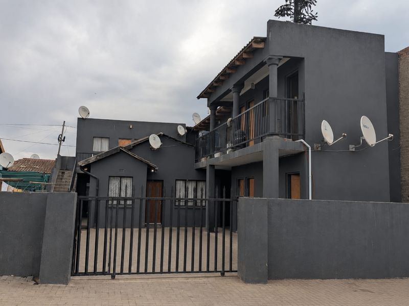 Front view of a multi-story apartment building featuring a paved driveway, secure metal gate, and balcony access.