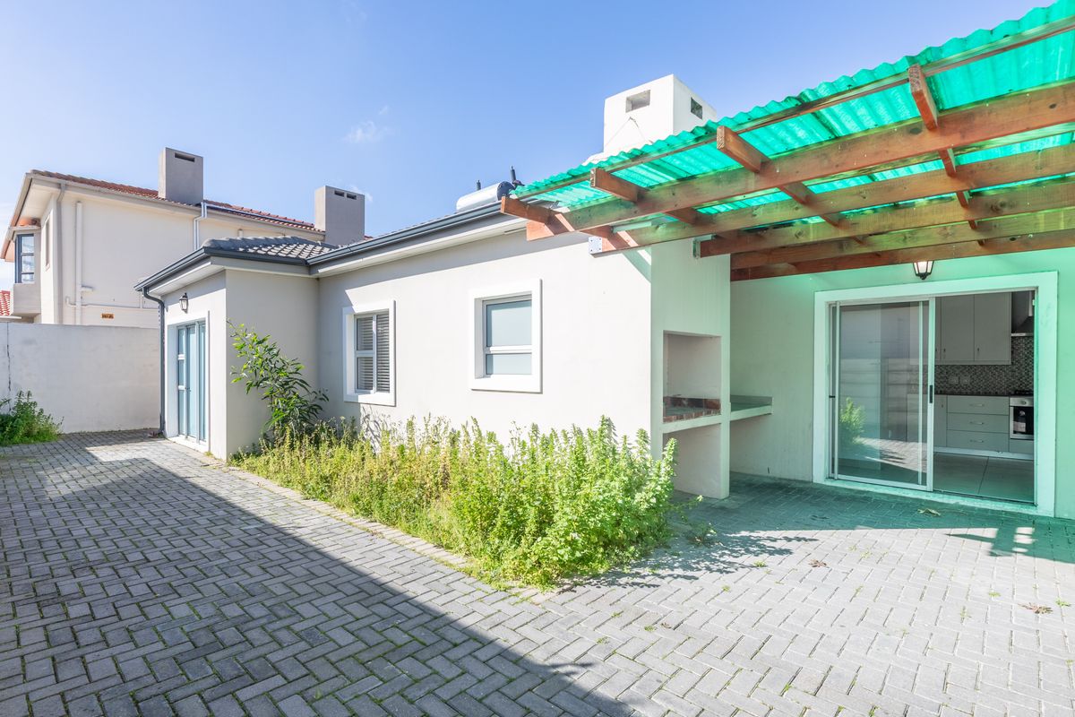 Rear exterior view featuring a paved patio area, built-in braai, and a timber pergola with green roofing, providing access to the home via sliding doors.