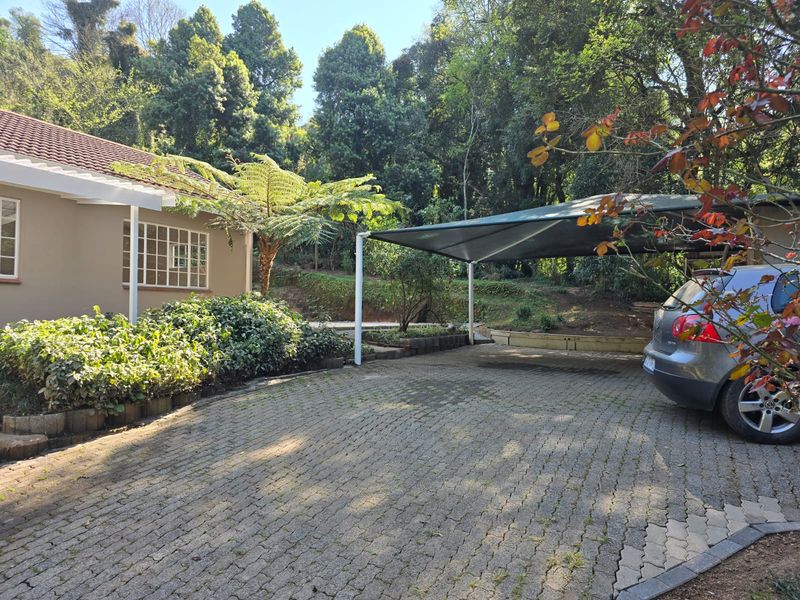 Front exterior view featuring a paved driveway, a shade cloth carport, and established garden surroundings with a tiled roof house facade.