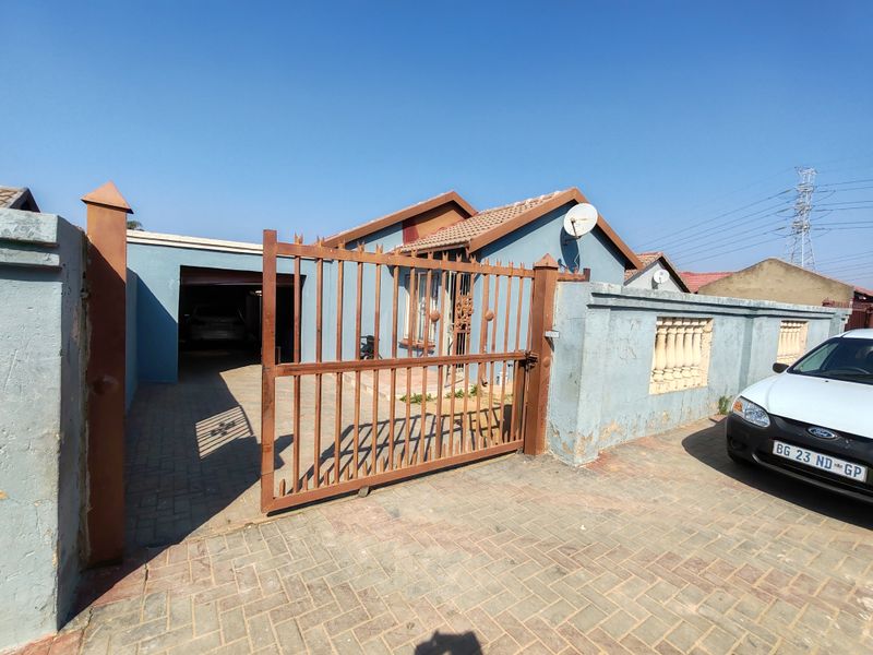 Front view of a residential property featuring a paved driveway, metal security gate, and perimeter wall with tiled roof structure.