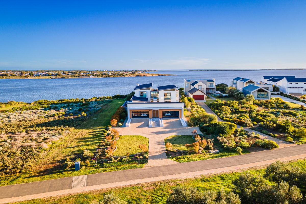 Aerial view of a modern waterfront residence featuring a double garage, paved driveway, and landscaped grounds in a scenic coastal setting.