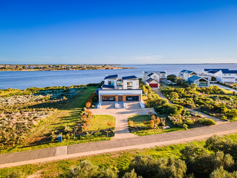 Aerial view of a modern waterfront residence featuring a double garage, paved driveway, and landscaped grounds in a scenic coastal setting.