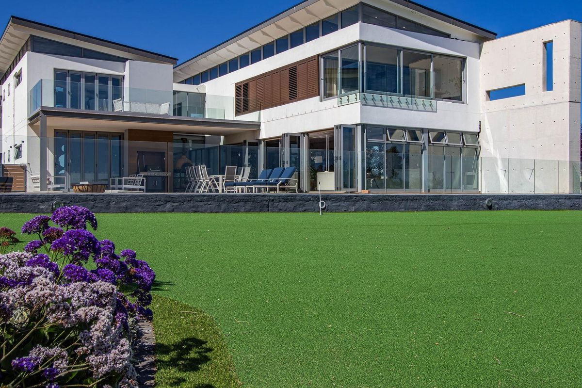 A modern rear elevation featuring expansive glass bi-fold doors, a covered patio area, and a well-maintained artificial lawn with landscaped garden beds.