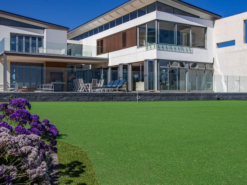 A modern rear elevation featuring expansive glass bi-fold doors, a covered patio area, and a well-maintained artificial lawn with landscaped garden beds.