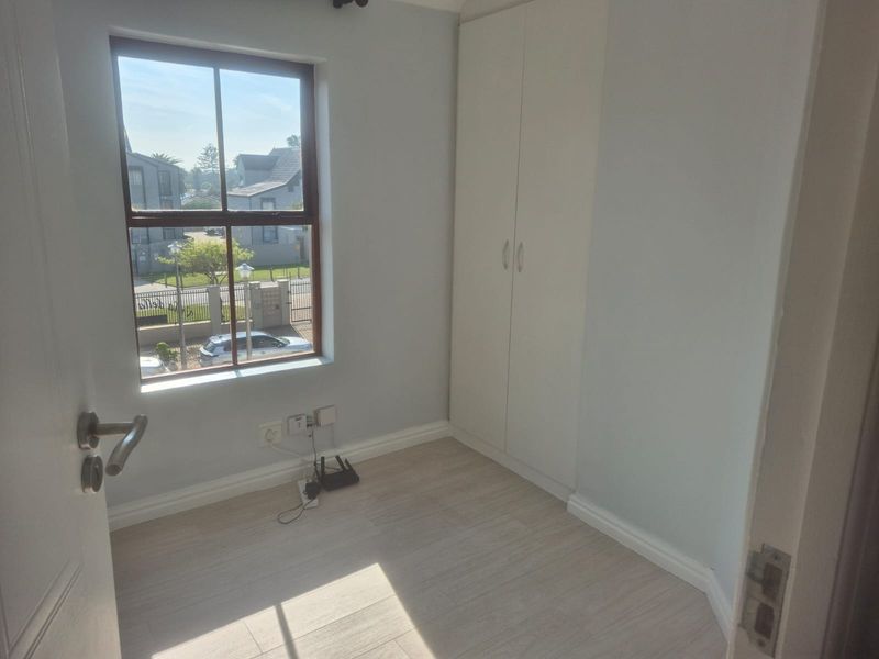 A bright, empty bedroom featuring light-toned laminate flooring, a built-in wardrobe, and a window overlooking the complex exterior.