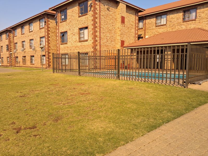 Communal swimming pool area featuring metal security fencing, set within a brick apartment complex with landscaped lawn and paved pathways.