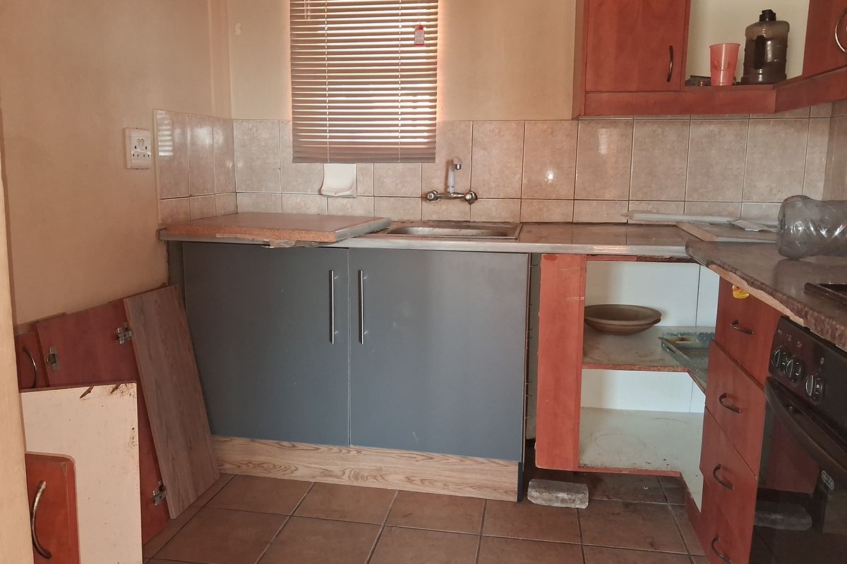 A dated kitchen featuring tiled flooring and backsplash, with significant damage to the built-in cabinetry and missing cabinet doors requiring repair or replacement.