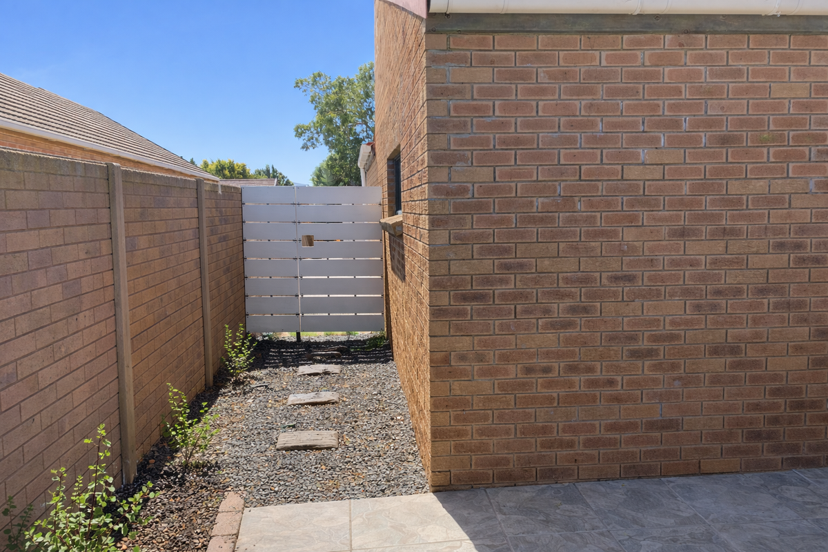A side passage of the cottage featuring a brick exterior wall, tiled patio section, stone pathway, and a modern horizontal slat gate.