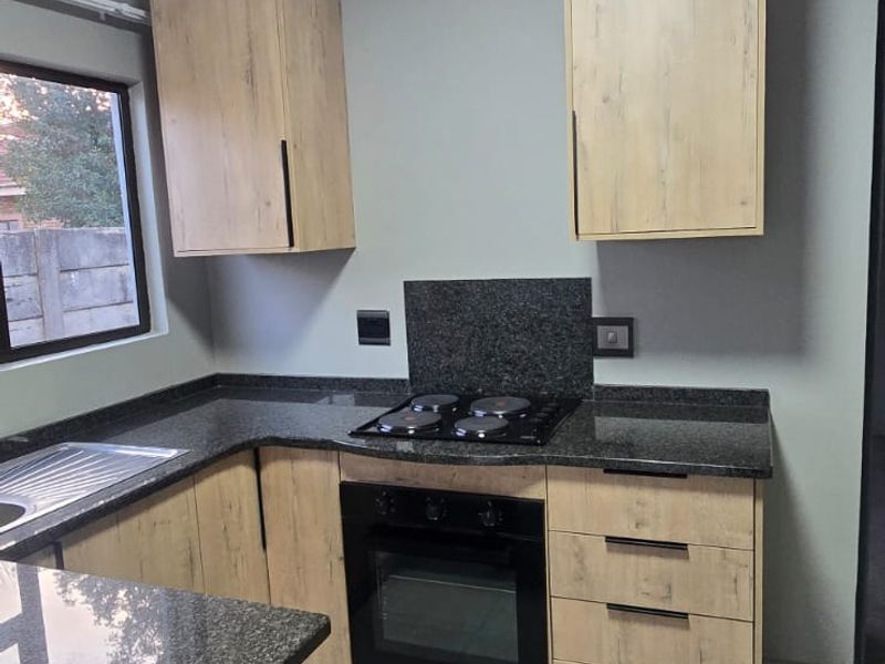 Modern kitchen featuring light wood-finish cabinetry, dark granite countertops, a built-in oven, electric hob, and neutral tiled flooring.