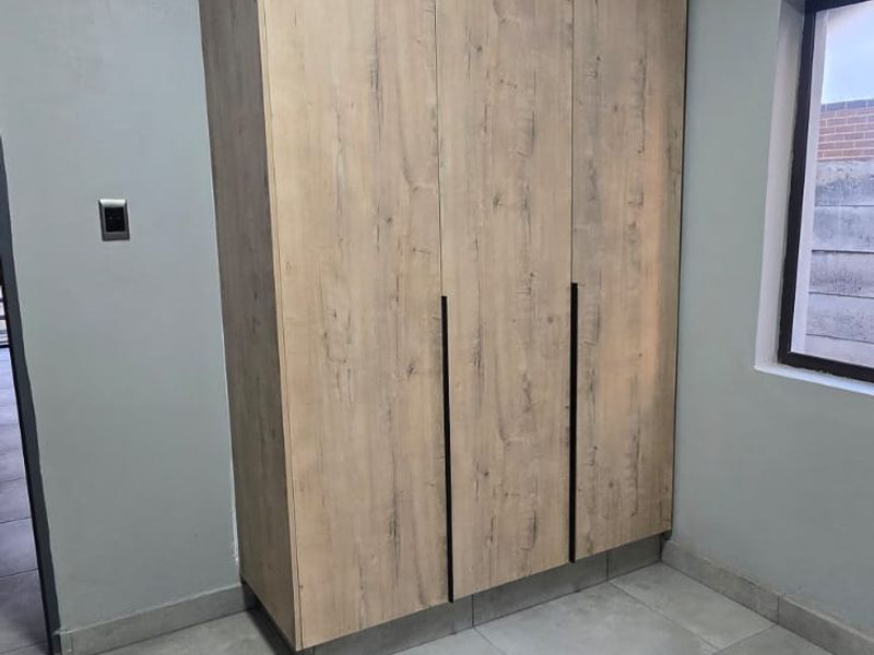 A clean, empty bedroom featuring modern wood-look built-in cupboards, neutral grey tiled flooring, and crisp white cornicing.