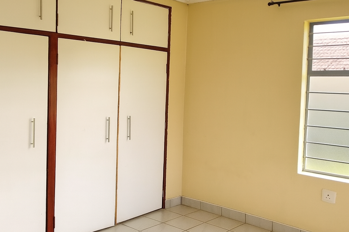 A clean, empty bedroom featuring tiled flooring, built-in cupboards with overhead storage, and a louvered window with a curtain rod.