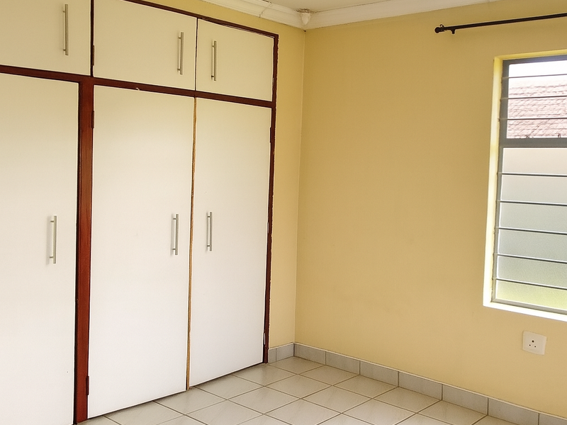 A clean, empty bedroom featuring tiled flooring, built-in cupboards with overhead storage, and a louvered window with a curtain rod.