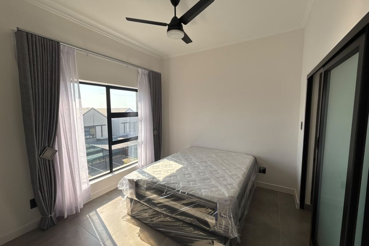 A bright bedroom featuring tiled flooring, a modern ceiling fan, and large windows with curtains, offering a clean and neutral living space.