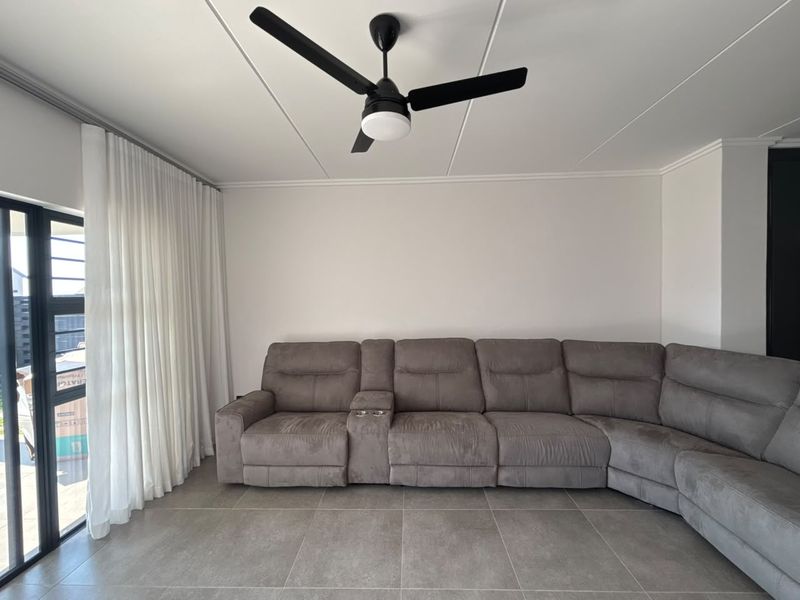 A modern living room featuring large format floor tiles, a contemporary ceiling fan, and a sliding glass door with floor-to-ceiling curtains.
