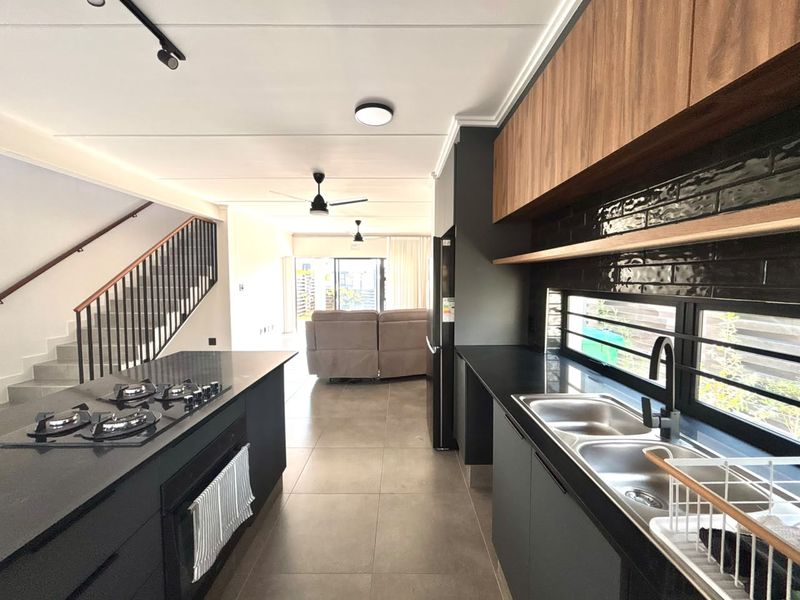 Modern open-plan kitchen featuring sleek dark cabinetry, gas hob, built-in oven, and double sink, seamlessly flowing into the living area with staircase access.