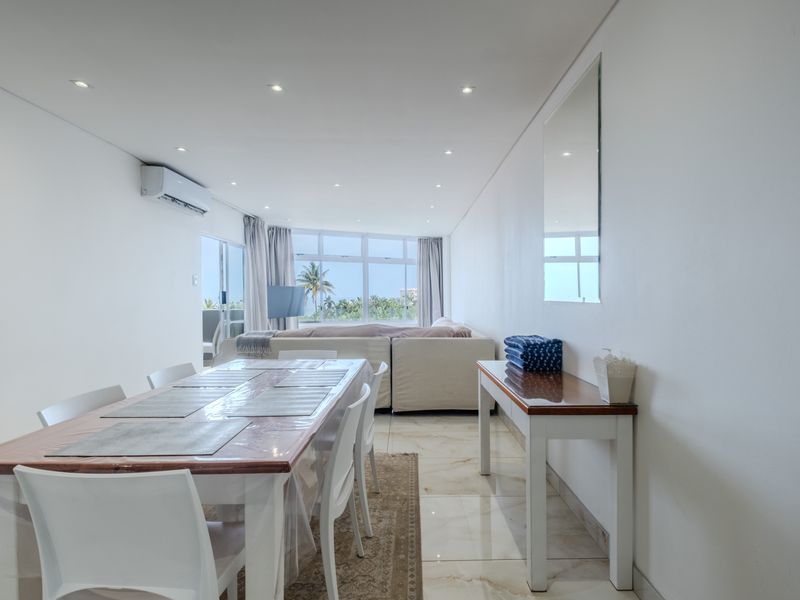 Bright open-plan living and dining area featuring polished tiled floors, recessed lighting, and a wall-mounted air conditioning unit with access to a balcony.
