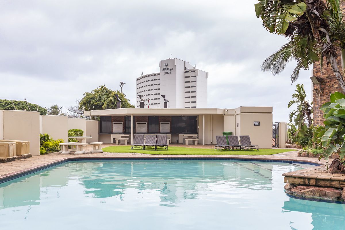 A well-maintained communal pool area featuring paved surrounds, built-in braai facilities, and established landscaping, set against a backdrop of local high-rise buildings.