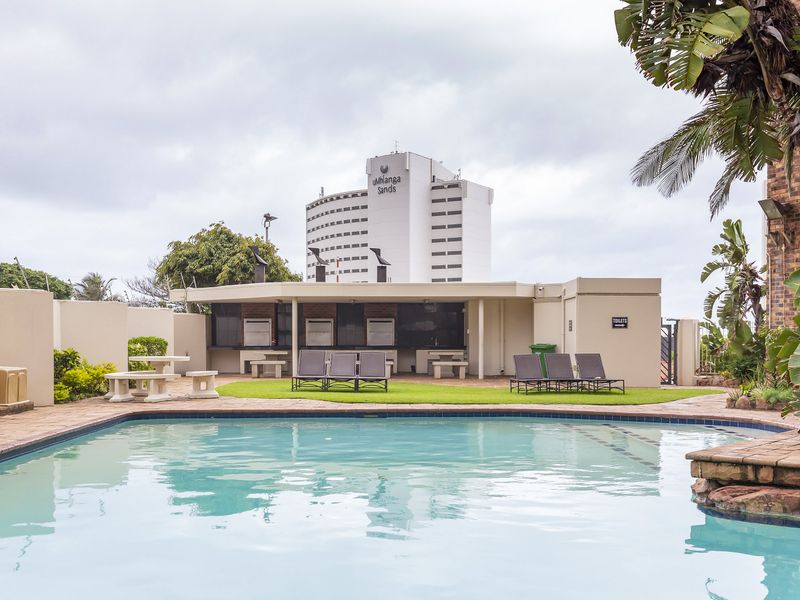 A well-maintained communal pool area featuring paved surrounds, built-in braai facilities, and established landscaping, set against a backdrop of local high-rise buildings.