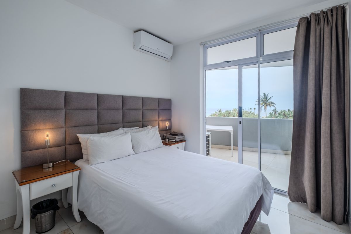 A bright bedroom featuring tiled floors, a wall-mounted air conditioning unit, and large sliding glass doors providing direct access to a private balcony.